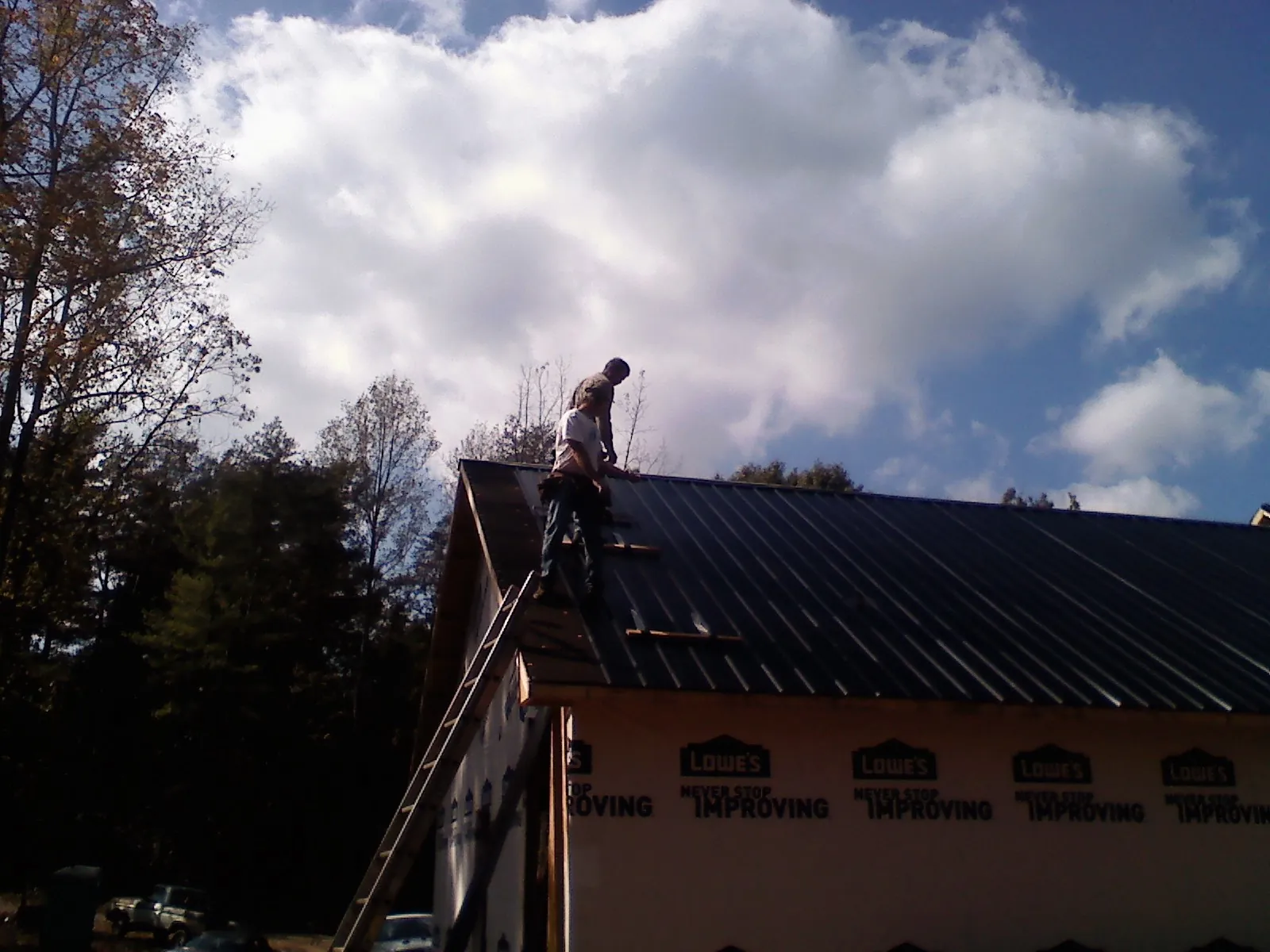Roofer fastening dark metal panels high against a cloudy sky, from a Roof Inspection job in Mastic
